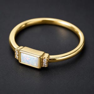 Puede incluir: Un anillo de oro con una piedra central de ópalo rectangular engastada horizontalmente. El engaste está flanqueado a ambos lados por dos pequeñas piedras redondas y claras. La banda es de oro liso y pulido.