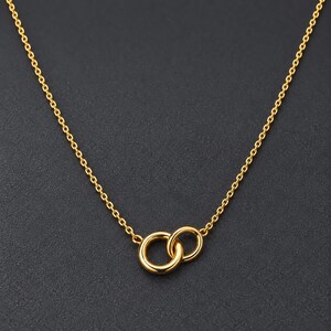 Può includere: Una collana color oro caratterizzata da una catenina sottile e un ciondolo composto da due anelli circolari intrecciati. Il metallo ha una finitura lucida e riflettente, e il design è centrato sulla catena, creando un aspetto semplice e simmetrico.