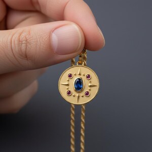 May include: Gold-colored pendant necklace with a circular pendant. The pendant features a central oval blue gemstone, surrounded by smaller red gemstones and starburst designs. The necklace chain is also gold-colored.