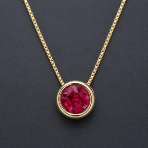 May include: A gold necklace featuring a round, faceted, deep red gemstone set in a smooth, polished gold bezel. The pendant is suspended from a delicate gold box chain, creating a refined and classic piece of jewelry with a vibrant center stone.