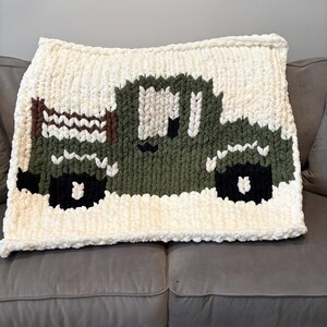 May include: A chunky knit blanket featuring a green truck design on a cream background. The truck has black wheels and a brown bed. The blanket is made of soft, thick yarn and is draped over a gray sofa.