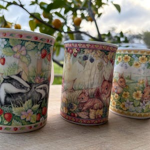 May include: Three ceramic mugs featuring detailed illustrations of woodland wildlife. The first mug displays a badger, the second shows squirrels with blackberries, and the third depicts rabbits among yellow flowers, all with decorative floral borders.