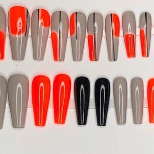 May include: A set of coffin-shaped false nails in various designs. The nails feature a combination of gray, bright orange, and black colors. Some nails have a vertical black stripe or orange accent.