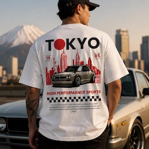 May include: A white short-sleeve t-shirt featuring a graphic on the back with the text TOKYO 1985, a red silhouette of a city skyline, a silver sports car, the words HIGH PERFORMANCE SPORTS, and a black and white checkered pattern at the bottom.