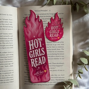 May include: A tall rectangular bookmark and a smaller circular sticker, both featuring a pink flame design and the text "HOT GIRLS READ with love" with a small yellow heart. The items are placed on the open pages of a book with printed text.