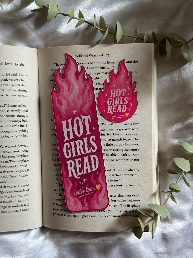 May include: A tall rectangular bookmark and a smaller circular sticker, both featuring a pink flame design and the text "HOT GIRLS READ with love" with a small yellow heart. The items are placed on the open pages of a book with printed text.