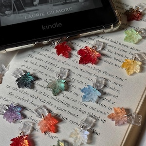 May include: A collection of small, translucent maple leaf-shaped charms in various colours, including red, orange, yellow, green, blue, and purple. Each charm features a clear plastic clip attachment and is scattered across an open book page.