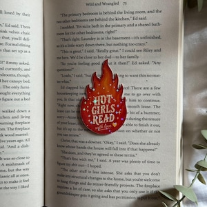 May include: A flame-shaped enamel pin with a red and orange gradient background, featuring the white text "HOT GIRLS READ" with small star and heart accents. The pin is positioned on the open pages of a book with printed text.