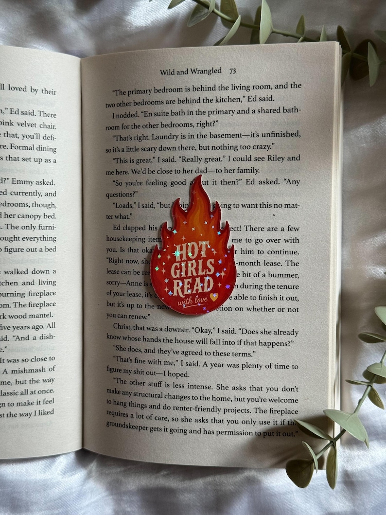May include: A flame-shaped enamel pin with a red and orange gradient background, featuring the white text "HOT GIRLS READ" with small star and heart accents. The pin is positioned on the open pages of a book with printed text.