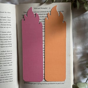 May include: Two flame-shaped bookmarks resting on an open book. One bookmark is a solid matte pink, and the other is a solid matte orange. Both bookmarks feature a stylized flame design at the top with rounded edges and a rectangular base.