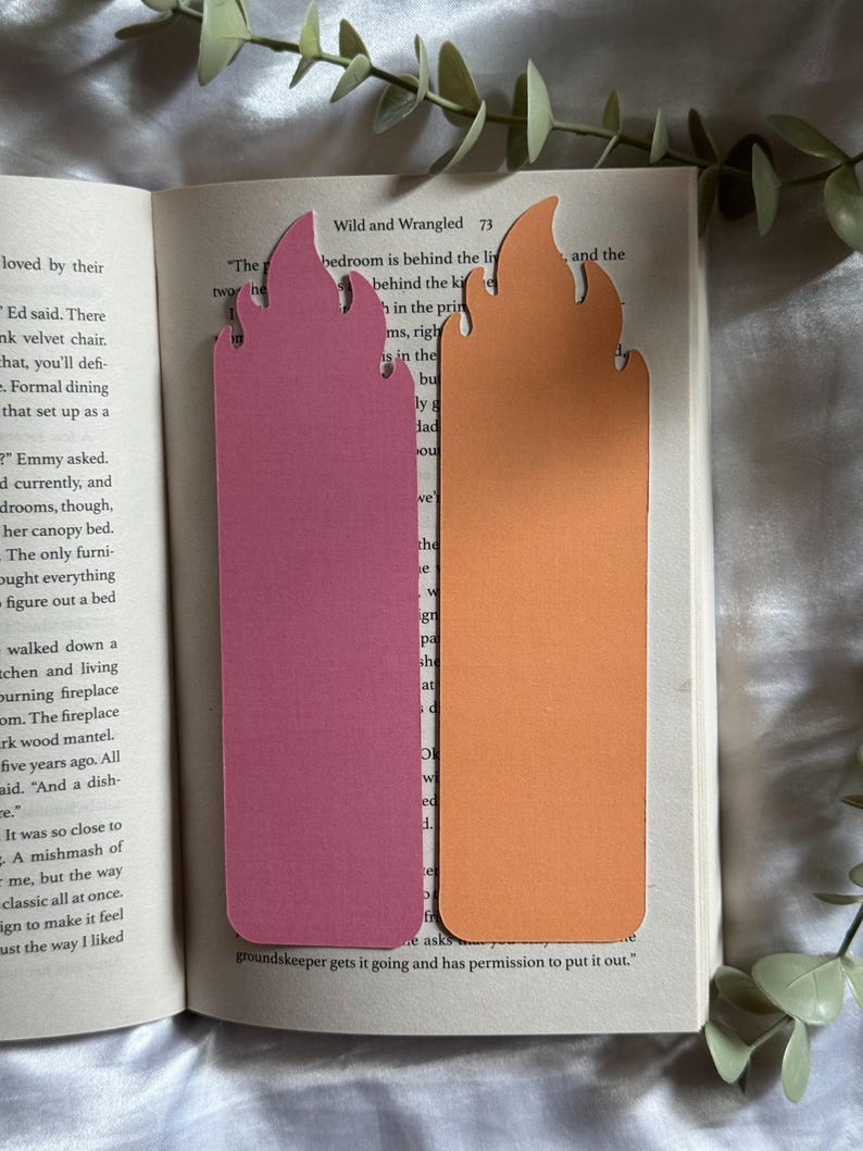 May include: Two flame-shaped bookmarks resting on an open book. One bookmark is a solid matte pink, and the other is a solid matte orange. Both bookmarks feature a stylized flame design at the top with rounded edges and a rectangular base.