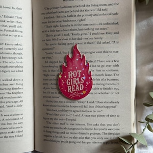 May include: A pink flame-shaped bookmark featuring the text "HOT GIRLS READ" in white lettering, accented with small white stars and a yellow heart at the bottom. The bookmark is placed on the open pages of a book with printed text.