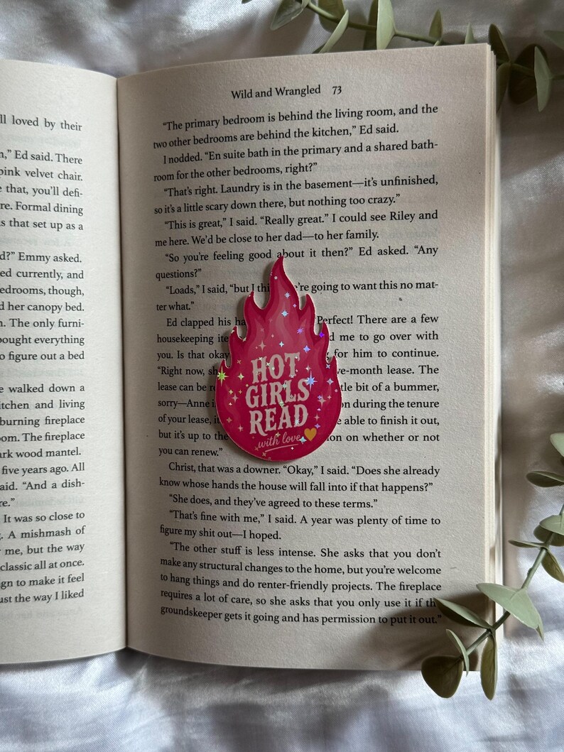 May include: A pink flame-shaped bookmark featuring the text "HOT GIRLS READ" in white lettering, accented with small white stars and a yellow heart at the bottom. The bookmark is placed on the open pages of a book with printed text.
