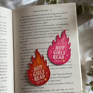May include: Two flame-shaped stickers placed on an open book page. The top sticker is pink with the white text "HOT GIRLS READ" and a small yellow heart. The bottom sticker is orange and red with the same white text and a yellow heart.