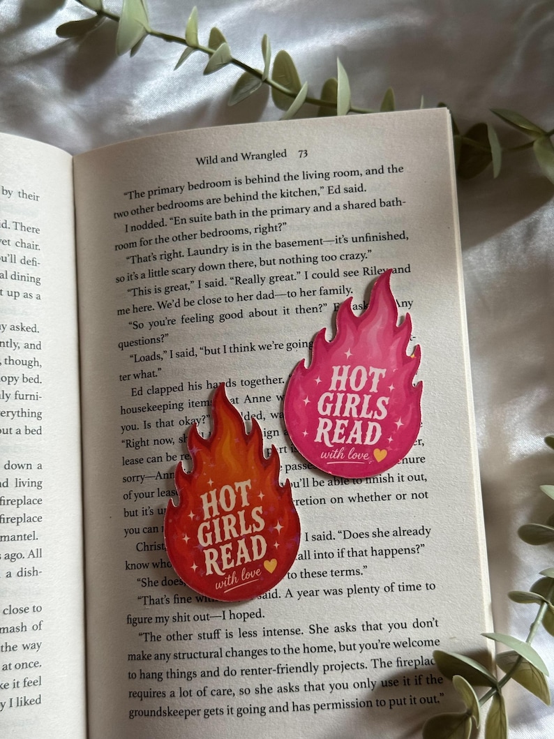 May include: Two flame-shaped stickers placed on an open book page. The top sticker is pink with the white text "HOT GIRLS READ" and a small yellow heart. The bottom sticker is orange and red with the same white text and a yellow heart.