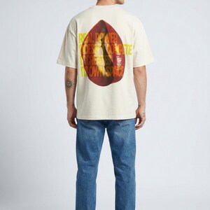Könnte beinhalten: Cremefarbenes T-Shirt mit großem, abstraktem Design auf dem Rücken. Das Design ist eine Kombination aus Rot und Orange mit gelbem Text. Das Model trägt blaue Jeans und weiße Turnschuhe.