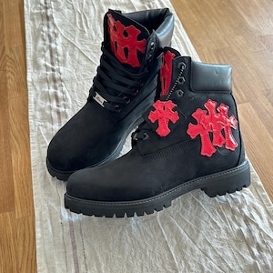 May include: A pair of black boots with red decorative accents. The boots have a classic lace-up design and a sturdy sole. The red accents are in a unique, stylized design. The boots are displayed on a neutral-colored fabric.