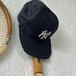 Peut inclure: Casquette de baseball noire avec un logo blanc. La casquette est présentée sur une raquette de tennis en bois vintage. La casquette a une visière incurvée et une couronne structurée. La raquette a un manche en bois et une poignée verte.