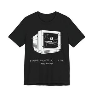 May include: Black t-shirt with a white graphic of a vintage computer monitor, displaying a cracked screen and a loading bar. The text "STATUS: PROCESSING... LIFE NOT FOUND" is printed below the monitor.