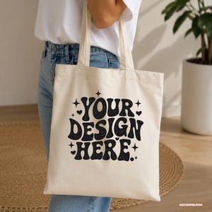 May include: A cream-colored canvas tote bag featuring the black text "YOUR DESIGN HERE" in a bold, wavy font. The design is accented with small black stars and heart shapes surrounding the lettering, which has a subtle speckled texture.