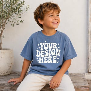 May include: A blue short-sleeved t-shirt featuring the white text "YOUR DESIGN HERE" in a wavy, retro-inspired font, accented with small star and heart graphics. The shirt has a crew neckline and a relaxed fit, displayed on a person.