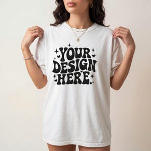 Może przedstawiać: Biała koszulka z napisem "YOUR DESIGN HERE" w czarnym, retro kroju pisma. Projekt zawiera gwiazdy i serca. Koszulka jest z krótkim rękawem i okrągłym dekoltem.
