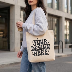 Puede incluir: Un bolso tote beige con el texto "YOUR DESIGN HERE" en letras negras de estilo retro. El bolso se lleva al hombro. La persona lleva una sudadera gris y vaqueros azules.