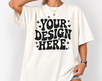 May include: An off-white short-sleeve t-shirt featuring black text that reads YOUR DESIGN HERE in a wavy, retro-inspired font. The design is accented with small black stars and hearts scattered around the lettering, creating a bold graphic.