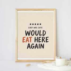 Op de afbeelding: Een ingelijste print met de tekst "Chef Was Cute, Would Eat Here Again" in zwart en rood. De print zit in een licht houten frame en staat op een wit oppervlak. Een kaars in een glazen pot staat naast het frame.