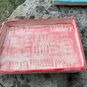 May include: A rectangular ceramic tray with a red rim and a textured, patterned interior. The tray's surface features a repeating oval design in a lighter shade of red, creating a contrast with the darker red border. The tray is resting on a grey stone surface.