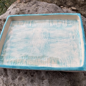 May include: Rectangular ceramic tray with a light blue rim and a white interior with a blue pattern. The tray has a handmade appearance, with visible texture and a glossy finish. The design is simple and elegant, suitable for serving or display.