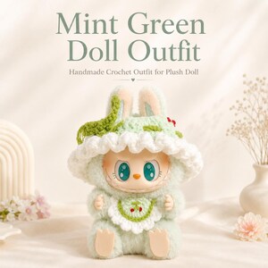 May include: A plush doll wearing a mint green crochet outfit, including a wide-brimmed hat with white trim and a bib featuring a small cherry graphic. The doll has large teal eyes, a small orange nose, and a wide grin. Text reads Mint Green Doll Outfit.