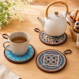 May include: A cream-coloured teapot with a bamboo handle rests on a floral coaster. A cup of tea sits on a blue floral coaster. Two additional coasters with decorative patterns are also visible. The coasters have a brown rope trim.