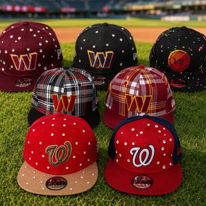 Op de afbeelding: Zeven baseballpetten met de letter "W" in verschillende kleuren en ontwerpen. Sommige petten zijn versierd met strass-steentjes. De petten worden getoond op een groen grasveld, wat wijst op een sport- of teamthema.