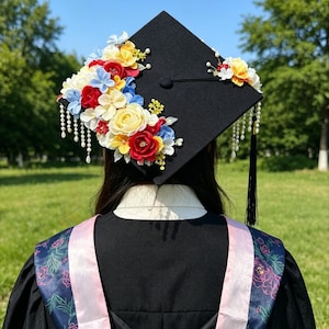 May include: A black mortarboard decorated with vibrant fabric flowers in red, yellow, blue, and white. The cap has a tassel and pearl-like bead embellishments. The person is wearing a black graduation gown with a pink and floral-patterned stole.