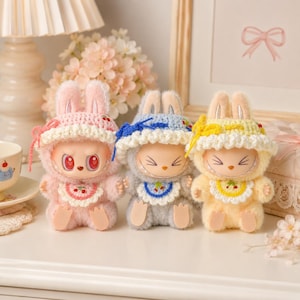 May include: Three small plush figures wearing crocheted bunny hoods and bibs. The left figure is pink with red eyes, the center is blue with closed eyes, and the right is yellow with closed eyes. Each features a white ruffled trim and cherry motif.