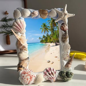 May include: A rectangular photo frame adorned with assorted seashells and a starfish. The frame has a white base with a light blue border. The image within shows a tropical beach with palm trees, white sand, and turquoise water.