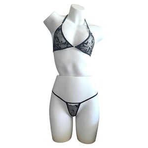 May include: A black lace lingerie set featuring a triangle bralette with thin halter straps and a matching thong. The fabric displays a detailed floral pattern in black lace over a sheer base, with thin black elastic bands forming the structure.
