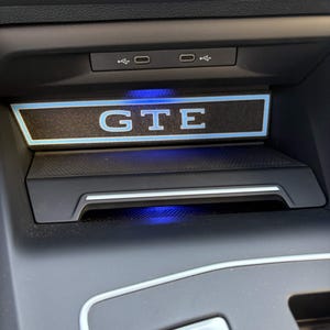 May include: A black automotive center console storage compartment featuring a backlit GTE logo in blue and white. The unit includes two USB-C ports above the compartment and a silver Engine Start/Stop button located below the storage area.