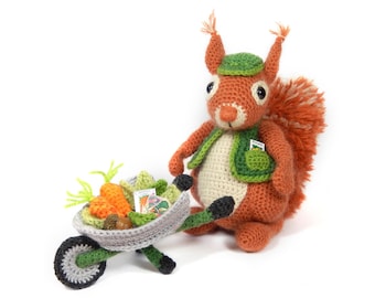 Peut inclure: Un écureuil en crochet portant un gilet vert et un chapeau, tenant une petite brouette verte remplie de carottes et de sachets de graines.
