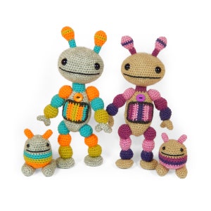 Puede incluir: Una familia de tres juguetes robot de crochet. Los dos robots más grandes tienen detalles en naranja, turquesa y morado. El robot más pequeño tiene detalles en naranja, turquesa y rosa.