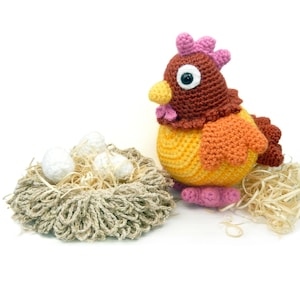 May include: A crocheted brown and yellow chicken with pink accents stands next to a nest filled with white crocheted eggs. The nest is made of brown yarn and is filled with straw.