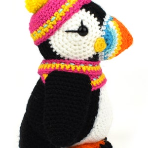 May include: A crocheted stuffed puffin wearing a pink and yellow hat with a yellow pom-pom and a pink and yellow scarf. The puffin has a white and black body with an orange beak and a black eye.