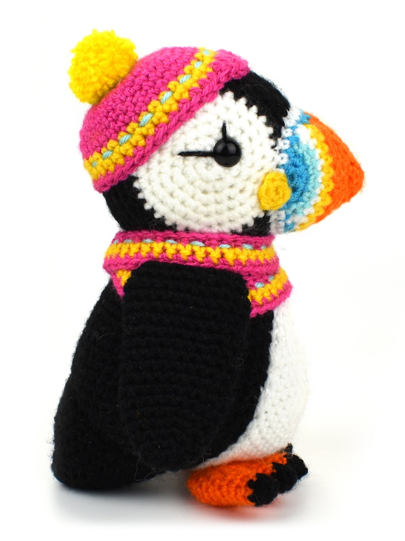 May include: A crocheted stuffed puffin wearing a pink and yellow hat with a yellow pom-pom and a pink and yellow scarf. The puffin has a white and black body with an orange beak and a black eye.