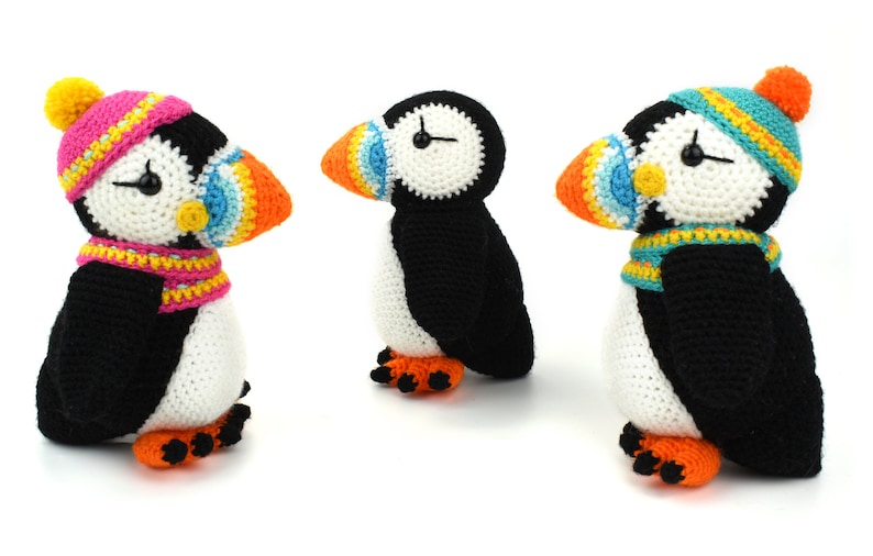 May include: Three crocheted stuffed puffins wearing colorful hats and scarves. The puffins are black and white with orange beaks and feet.