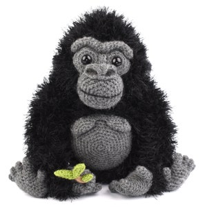 以下が含まれることがあります： 黒い毛皮と灰色の顔をした、かぎ針編みで作られたゴリラのぬいぐるみ。ゴリラは手に緑の葉を持っています。