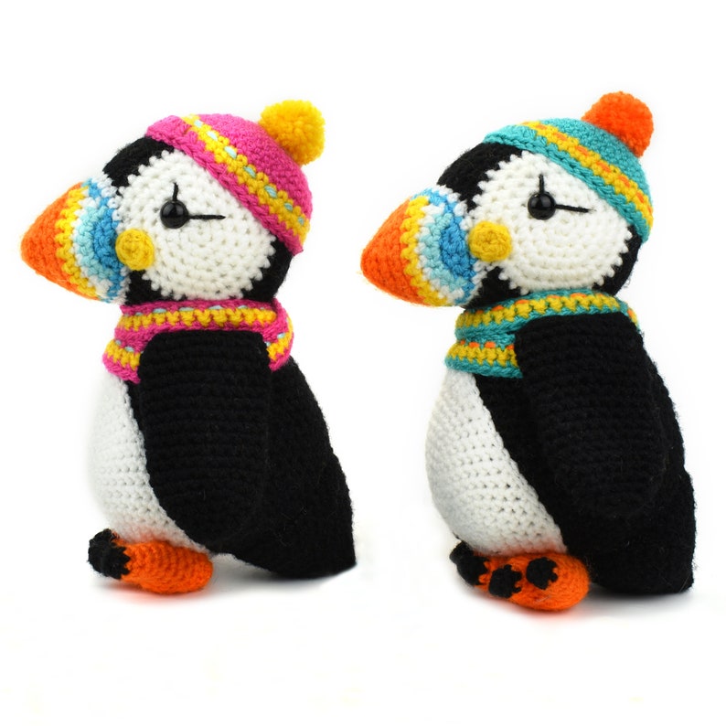 May include: Two crocheted stuffed puffins wearing colorful hats and scarves. The puffin on the left is wearing a pink and yellow hat and scarf, while the puffin on the right is wearing a teal and yellow hat and scarf.