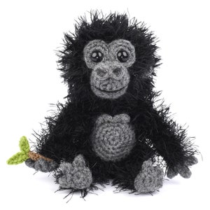 以下が含まれることがあります： 緑の葉っぱを持った黒い毛糸のゴリラのぬいぐるみが座っています。ぬいぐるみは灰色の顔と灰色の胸をしています。