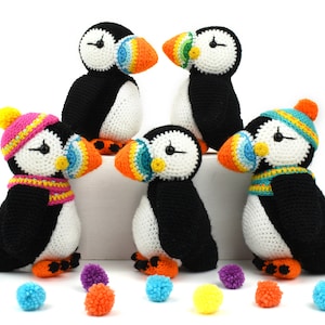 May include: Four crocheted stuffed puffins with colorful beaks and hats. The puffins are black and white with orange, yellow, blue, and green accents. The puffins are sitting on a white surface with colorful pom-poms scattered around them.