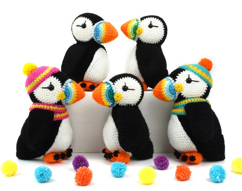 May include: Four crocheted stuffed puffins with colorful beaks and hats. The puffins are black and white with orange, yellow, blue, and green accents. The puffins are sitting on a white surface with colorful pom-poms scattered around them.